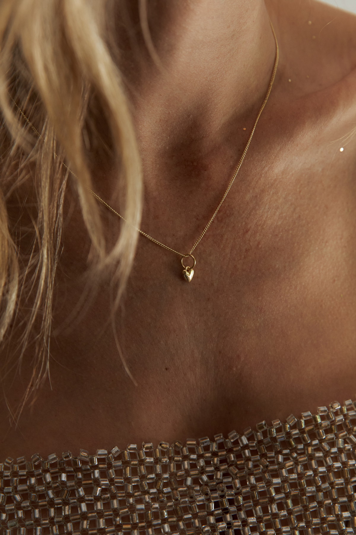 A close-up lifestyle shot of a model wearing a delicate gold chain necklace with a small heart-shaped pendant, paired with a sparkling champagne-colored beaded top.