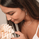 A woman sits with a flower wearing the You Are Loved Earrings in sterling silver. These earrings feature a small heart pendant that hangs elegantly from a hoop earring. The You Are Loved Earrings are part of the Revie Jane x Bluebird Co exclusive Mother's Day collection.