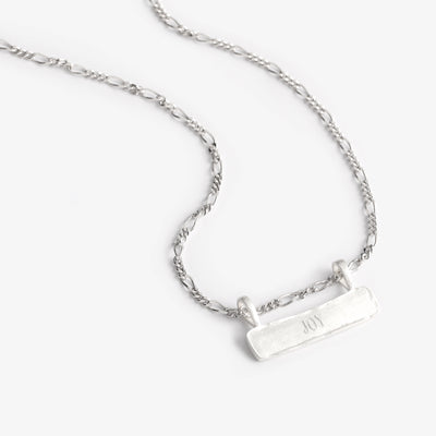 A close up look at the Signature Bar Necklace with a fine figaro chain and solid sterling silver bar pendant that has been engraved with the word Joy.